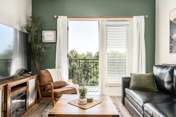 Living room with sage green walls, black leather sofa, brown leather armchair, wooden TV stand, and glass doors opening to a balcony with tree views.